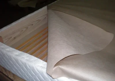 Pose du tissu d’habillage sur les lattes d’un sommier tapissier Ox Literie, entouré d’un coutil matelassé blanc