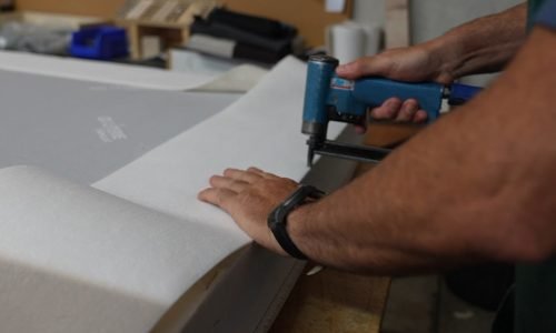Découpe et pose de la toile de recouvrement sur un sommier tapissier habillé dans l'atelier OX Literie