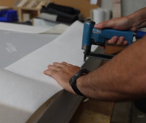 Découpe et pose de la toile de recouvrement sur un sommier tapissier habillé dans l'atelier OX Literie